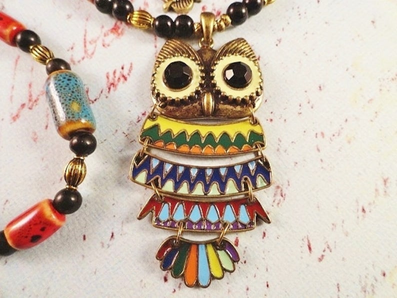 May include: A colorful owl pendant necklace with black beads and red and blue beads. The owl is made of metal and has a layered design with geometric patterns.