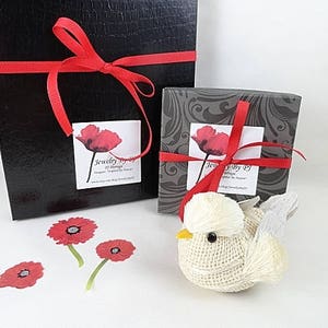 May include: Two gift boxes, one black and one gray, are tied with red ribbons. Each box has a label with a red poppy design. A small, white bird figurine with a yellow beak sits nearby, along with three poppy stickers.