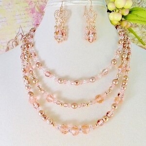 K&ouml;nnte beinhalten: Ein mehrreihiges Halsketten- und Ohrring-Set mit ros&eacute;goldenen und blush-rosa Perlen. Die Halskette hat vier Reihen Perlen, darunter kleine runde Perlen, gr&ouml;&szlig;ere runde Perlen und facettierte Perlen. Die Ohrringe sind H&auml;ngeohrringe mit einem herzf&ouml;rmigen Design.