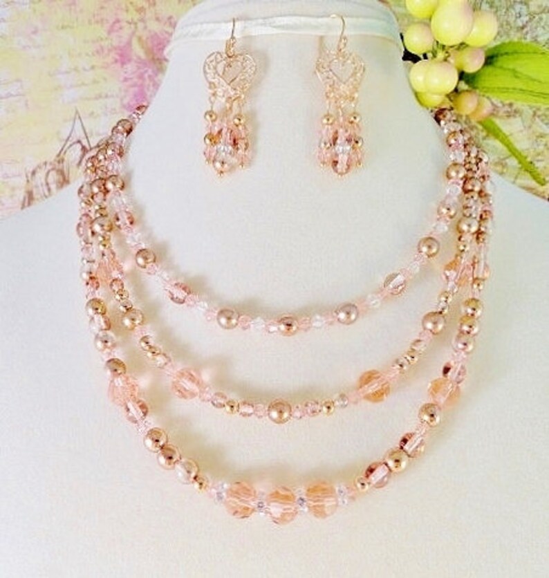 K&ouml;nnte beinhalten: Ein mehrreihiges Halsketten- und Ohrring-Set mit ros&eacute;goldenen und blush-rosa Perlen. Die Halskette hat vier Reihen Perlen, darunter kleine runde Perlen, gr&ouml;&szlig;ere runde Perlen und facettierte Perlen. Die Ohrringe sind H&auml;ngeohrringe mit einem herzf&ouml;rmigen Design.