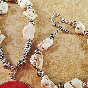 May include: A white howlite stone necklace with silver accents and a red coral pendant. The necklace has a lobster clasp closure.