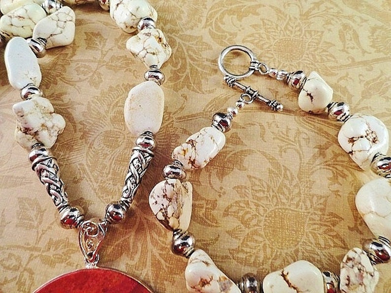 May include: A white howlite stone necklace with silver accents and a red coral pendant. The necklace has a lobster clasp closure.