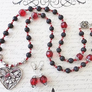 May include: A black lava rock and red crystal beaded necklace with a silver heart pendant and matching earrings. The heart pendant has a floral design.