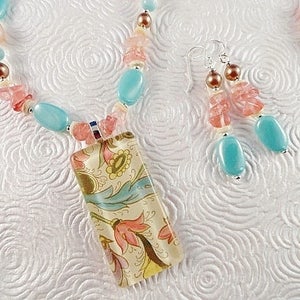 May include: A turquoise and pink beaded necklace with a floral patterned pendant. The necklace is made with various sized beads and has a silver clasp. The pendant is rectangular and has a floral design in shades of blue, pink, and yellow.