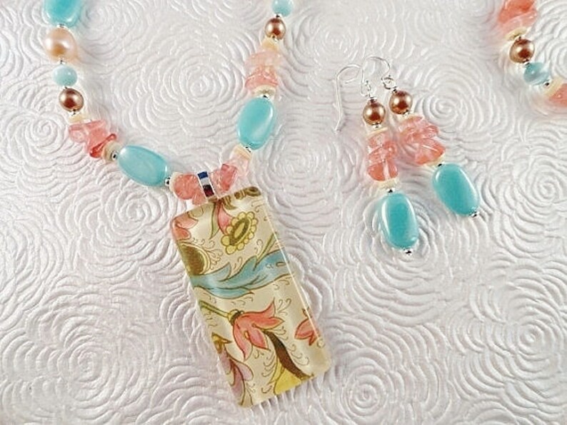 May include: A turquoise and pink beaded necklace with a floral patterned pendant. The necklace is made with various sized beads and has a silver clasp. The pendant is rectangular and has a floral design in shades of blue, pink, and yellow.