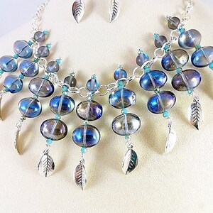 May include: A handcrafted necklace and earring set featuring iridescent blue and silver beads. The necklace has a silver chain with dangling leaf charms and turquoise accents. The earrings are small leaf-shaped studs.