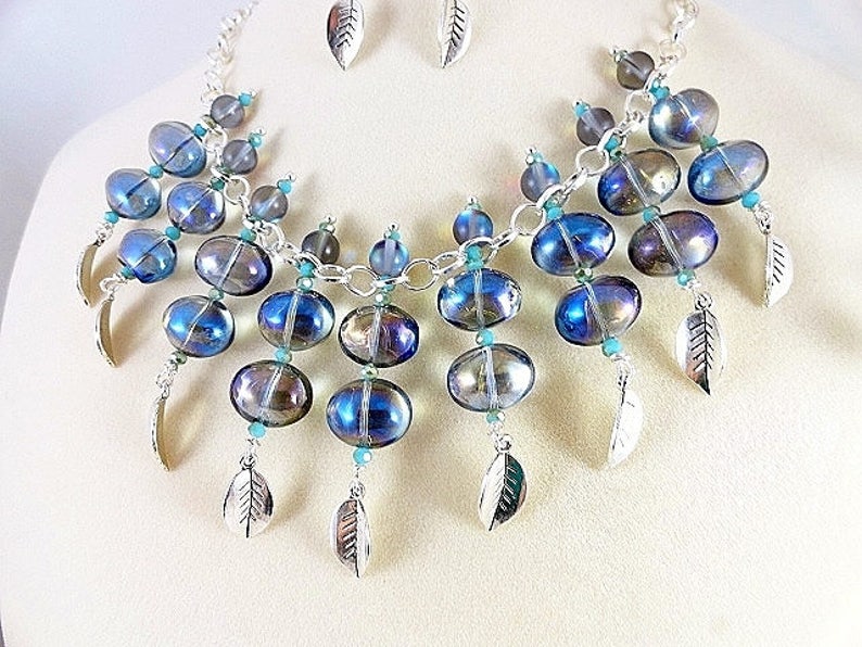 May include: A handcrafted necklace and earring set featuring iridescent blue and silver beads. The necklace has a silver chain with dangling leaf charms and turquoise accents. The earrings are small leaf-shaped studs.