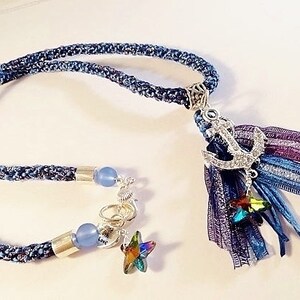 May include: A blue denim rope necklace with a silver anchor charm and a multi-colored tassel. The tassel is made of blue, purple, and silver ribbons. The necklace also has two blue glass beads and a silver star charm.