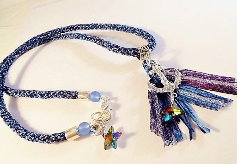 May include: A blue denim rope necklace with a silver anchor charm and a multi-colored tassel. The tassel is made of blue, purple, and silver ribbons. The necklace also has two blue glass beads and a silver star charm.