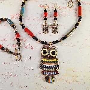 May include: A colorful owl pendant necklace set with black, red, and blue beads. The owl pendant has a multi-colored design with yellow, green, blue, and red accents. The set also includes a matching pair of owl earrings.