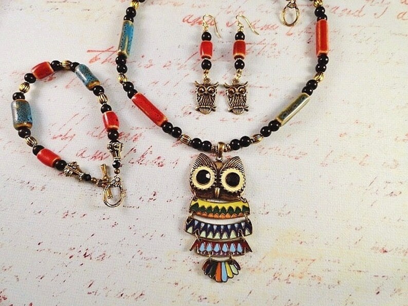 May include: A colorful owl pendant necklace set with black, red, and blue beads. The owl pendant has a multi-colored design with yellow, green, blue, and red accents. The set also includes a matching pair of owl earrings.