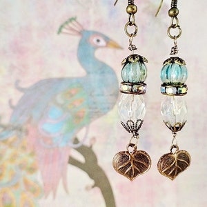 May include: A pair of dangle earrings with a blue and iridescent glass bead, a clear crystal bead, and a leaf-shaped charm. The earrings are made with bronze-colored metal.