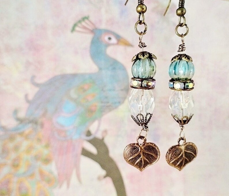 May include: A pair of dangle earrings with a blue and iridescent glass bead, a clear crystal bead, and a leaf-shaped charm. The earrings are made with bronze-colored metal.