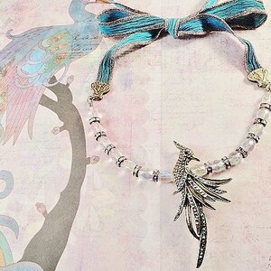 May include: A silver and iridescent crystal necklace with a silver bird pendant. The necklace is accented with a turquoise blue ribbon.