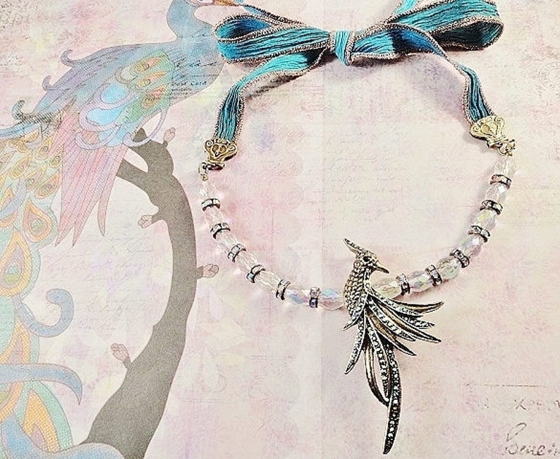 May include: A silver and iridescent crystal necklace with a silver bird pendant. The necklace is accented with a turquoise blue ribbon.