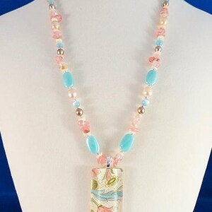 May include: A necklace with a rectangular pendant featuring a floral pattern in shades of pink, blue, and yellow. The necklace is made of pink, blue, and white beads.