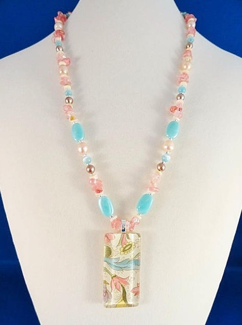 May include: A necklace with a rectangular pendant featuring a floral pattern in shades of pink, blue, and yellow. The necklace is made of pink, blue, and white beads.