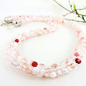 May include: A delicate necklace featuring a beaded design in shades of pink and white, accented with red beads. The necklace has a silver clasp and is displayed on a white surface. A sprig of flowers is in the background.