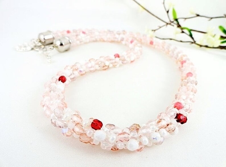 May include: A delicate necklace featuring a beaded design in shades of pink and white, accented with red beads. The necklace has a silver clasp and is displayed on a white surface. A sprig of flowers is in the background.