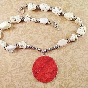 May include: A white howlite stone necklace with a large, round, red coral pendant. The necklace is made with silver beads and a silver clasp.