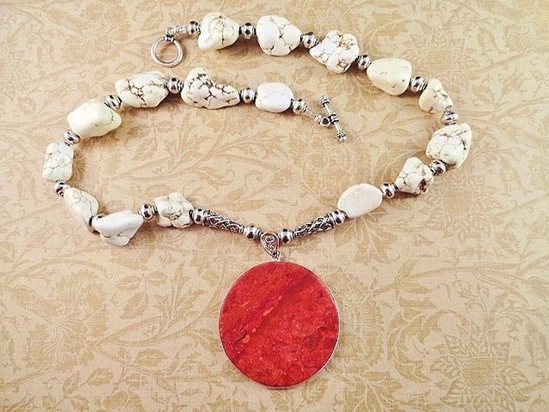 May include: A white howlite stone necklace with a large, round, red coral pendant. The necklace is made with silver beads and a silver clasp.