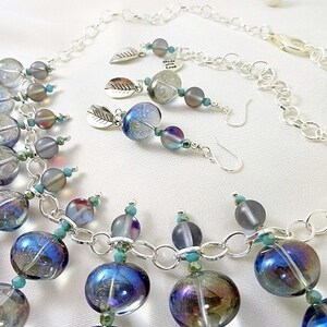 May include: A jewelry set featuring a necklace and earrings. The necklace has large, iridescent blue and clear glass beads connected by a silver chain. The earrings have similar beads and silver leaf charms. The set is on a white background.