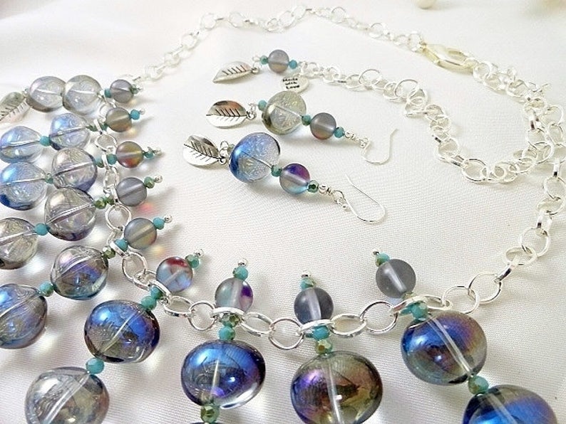 May include: A jewelry set featuring a necklace and earrings. The necklace has large, iridescent blue and clear glass beads connected by a silver chain. The earrings have similar beads and silver leaf charms. The set is on a white background.