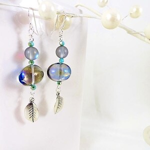 May include: Dangle earrings with iridescent glass beads in shades of blue, green, and gold. The earrings feature silver-tone leaf charms and are accented with teal and clear beads. The earrings hang from silver-tone ear wires.