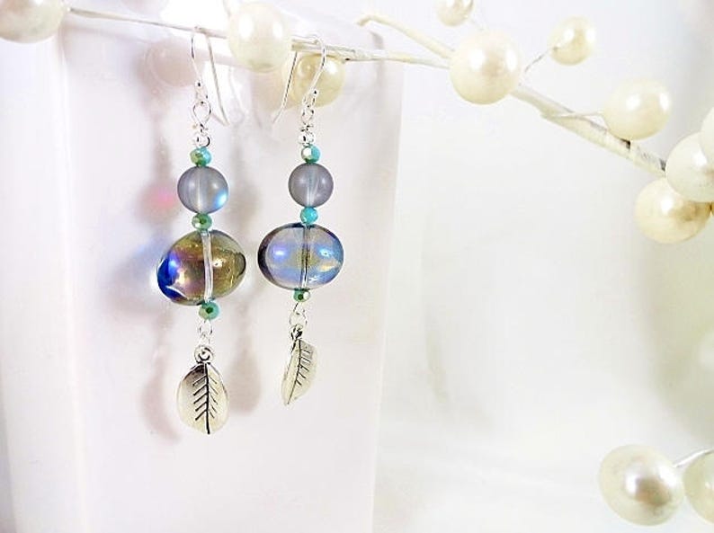 May include: Dangle earrings with iridescent glass beads in shades of blue, green, and gold. The earrings feature silver-tone leaf charms and are accented with teal and clear beads. The earrings hang from silver-tone ear wires.