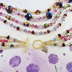 May include: Multi-strand necklace featuring gold beads and purple, pink, and clear crystals. The necklace has a gold toggle clasp and a small heart charm inscribed with the words "Made with Love".