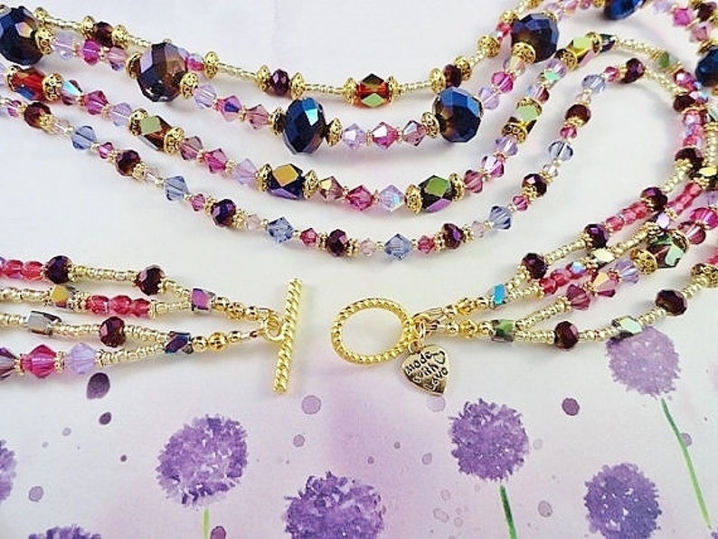 May include: Multi-strand necklace featuring gold beads and purple, pink, and clear crystals. The necklace has a gold toggle clasp and a small heart charm inscribed with the words "Made with Love".