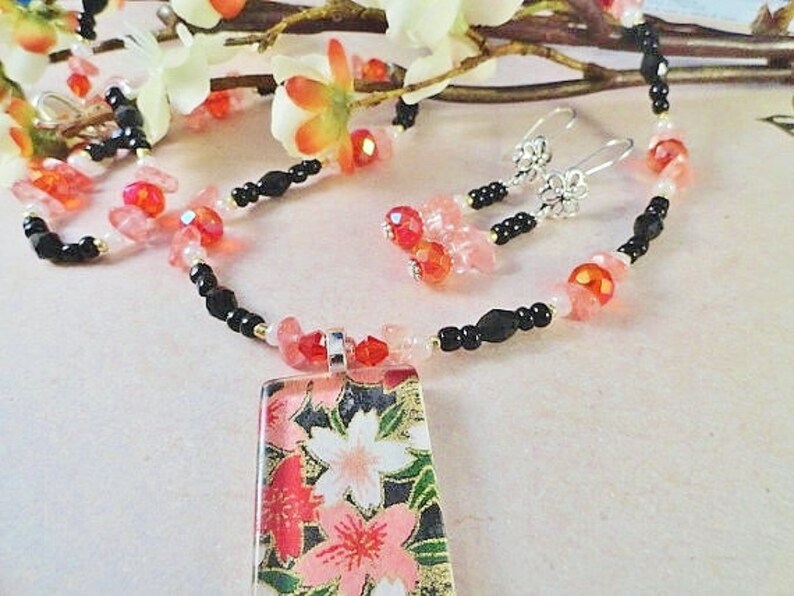May include: A delicate necklace with a rectangular pendant featuring a floral design. The necklace is made with black beads, pink and red glass beads, and a silver chain. The pendant is made of glass and has a floral pattern with pink and red flowers on a black background.