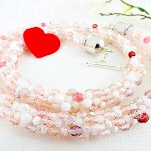 May include: A beaded necklace with a mix of pink, white, and clear beads. The necklace has a silver clasp and a red heart-shaped accent. The beads are arranged in a braided pattern, creating a delicate and feminine look.