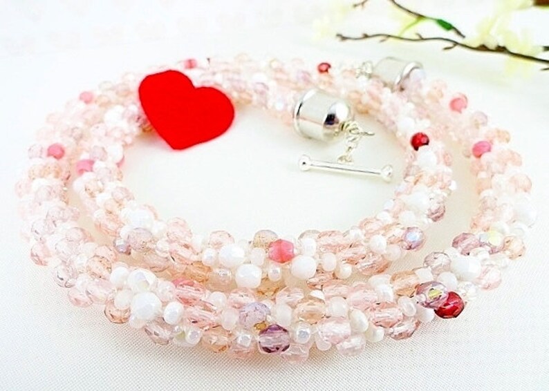May include: A beaded necklace with a mix of pink, white, and clear beads. The necklace has a silver clasp and a red heart-shaped accent. The beads are arranged in a braided pattern, creating a delicate and feminine look.