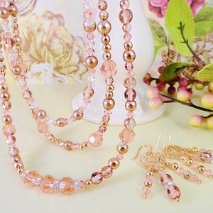 K&ouml;nnte beinhalten: Ein Set aus zarten rosafarbenen und goldenen Perlen-Schmuck, bestehend aus einer mehrlagigen Halskette und passenden Ohrringen. Die Halskette besteht aus mehreren Str&auml;ngen kleiner Perlen in Rosat&ouml;nen und Gold, w&auml;hrend die Ohrringe schlicht und elegant sind, mit einem einzigen Tropfen aus rosafarbenen Perlen.
