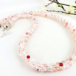 May include: A delicate necklace composed of small, light pink and clear beads, interspersed with a few red beads. The necklace has a silver clasp and a toggle closure. The beads are arranged in a twisted pattern.