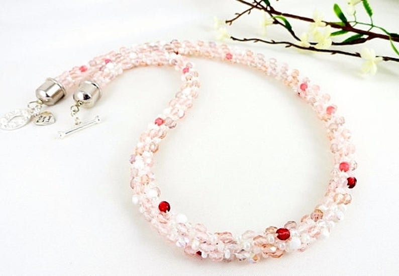 May include: A delicate necklace composed of small, light pink and clear beads, interspersed with a few red beads. The necklace has a silver clasp and a toggle closure. The beads are arranged in a twisted pattern.