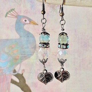 May include: A pair of dangle earrings with a light blue and clear glass bead, a silver charm, and a leaf-shaped pendant. The earrings are on a white background with a blurred image of a peacock.