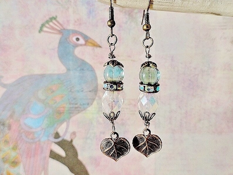 May include: A pair of dangle earrings with a light blue and clear glass bead, a silver charm, and a leaf-shaped pendant. The earrings are on a white background with a blurred image of a peacock.