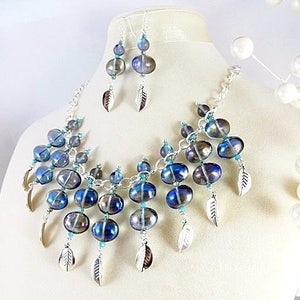 May include: A necklace and earring set featuring iridescent blue and silver beads. The necklace and earrings have silver feather charms. The jewelry is displayed on a mannequin.