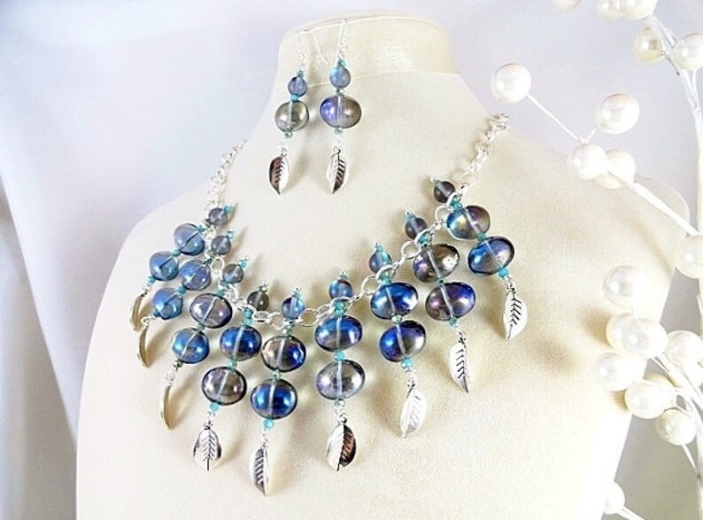 May include: A necklace and earring set featuring iridescent blue and silver beads. The necklace and earrings have silver feather charms. The jewelry is displayed on a mannequin.