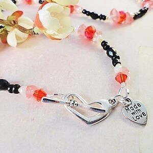 May include: A delicate beaded necklace with black, pink, and white beads. The necklace has a silver heart charm with the words "Made with Love" engraved on it.