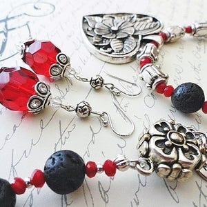 May include: A silver necklace with a floral heart pendant, red and black beads, and a smaller silver charm with a flower design. The necklace comes with matching earrings with red glass beads and silver accents. The charm on the necklace has the text "Made with Love".