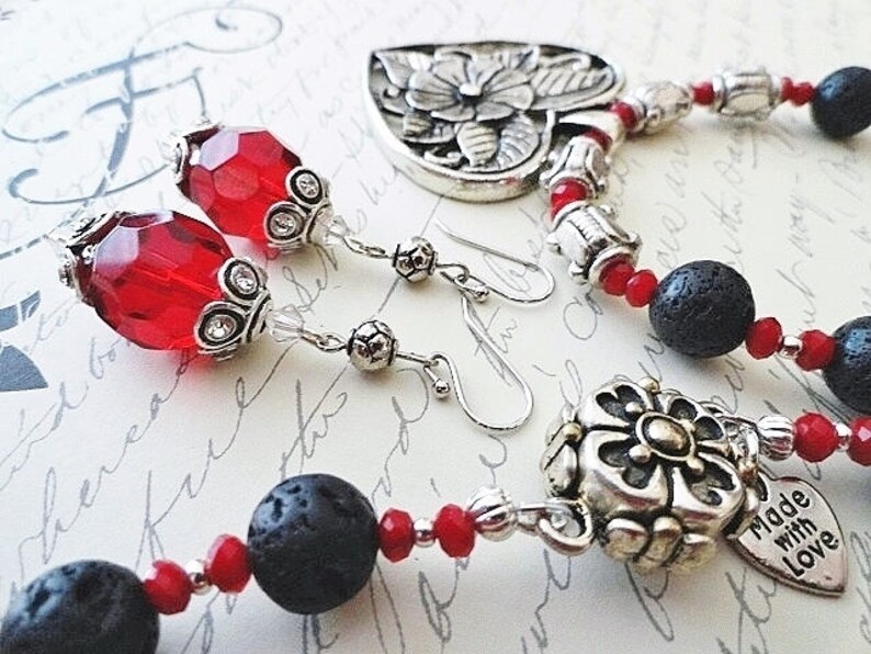 May include: A silver necklace with a floral heart pendant, red and black beads, and a smaller silver charm with a flower design. The necklace comes with matching earrings with red glass beads and silver accents. The charm on the necklace has the text "Made with Love".