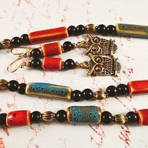 May include: A necklace and earrings set featuring black, red, and blue ceramic beads, gold accents, and owl charms.
