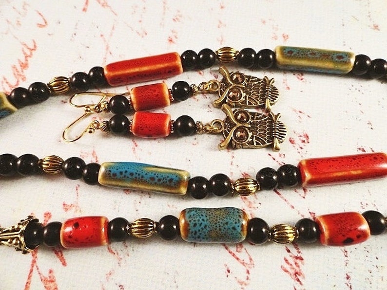 May include: A necklace and earrings set featuring black, red, and blue ceramic beads, gold accents, and owl charms.