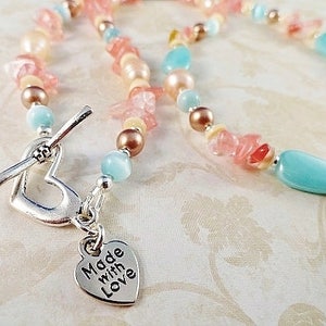 May include: A delicate necklace made with pastel pink, blue, and white beads. The necklace features a heart-shaped clasp with a charm that reads "Made with Love".