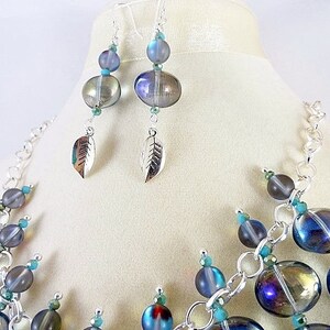 May include: A necklace and earring set featuring iridescent blue and gray glass beads. The necklace has a silver chain and turquoise accents. The earrings have leaf charms. The jewelry is displayed on a mannequin.