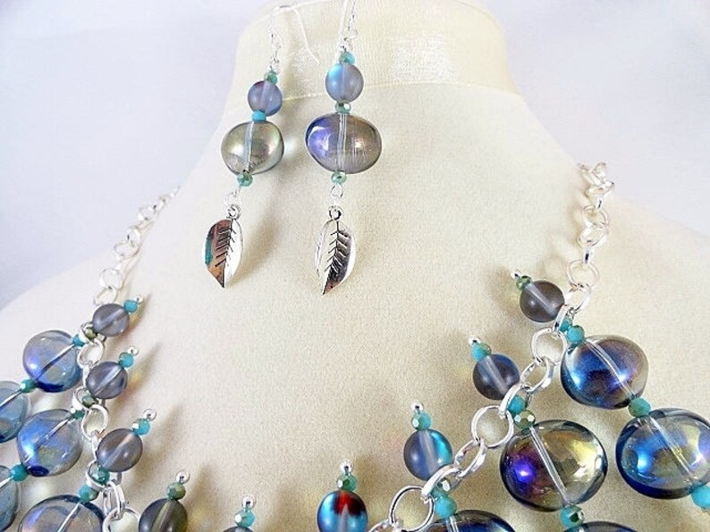 May include: A necklace and earring set featuring iridescent blue and gray glass beads. The necklace has a silver chain and turquoise accents. The earrings have leaf charms. The jewelry is displayed on a mannequin.