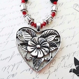 May include: A silver heart-shaped pendant necklace with a floral design. The necklace is made of red beads and silver beads.